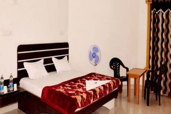A single bed with a red and white patterned bedspread, a white pillow, and a black headboard. There is a small wooden table and a black chair to the right of the bed. A wall-mounted fan is visible on the wall behind the bed. - Shiv Tara Desert Camp (Jaisalmer)