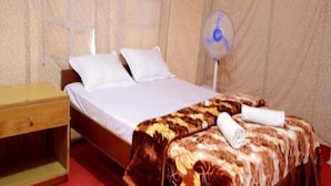 A single bed with white linens, a wooden nightstand, a patterned blanket, and a standing fan. - Shiv Tara Desert Camp (Jaisalmer)