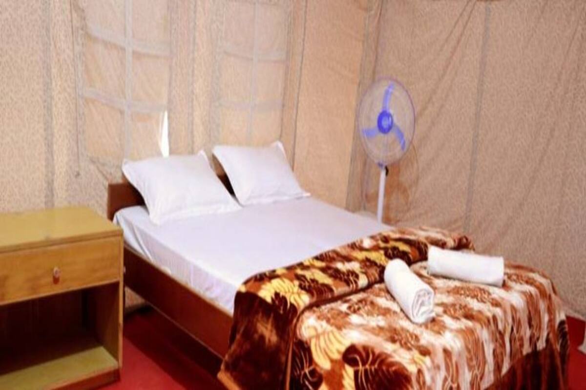 A single bed with white linens, a wooden nightstand, a patterned blanket, and a standing fan.