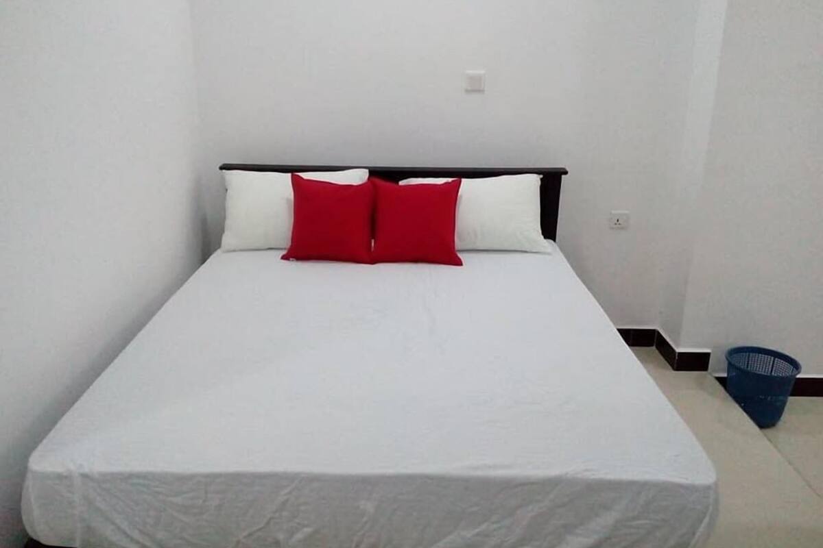 A single bed with white bedding and two red pillows, a blue basket, and a white wall.