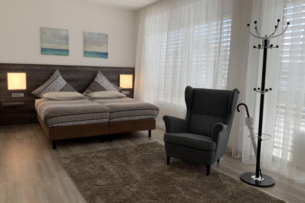 A bedroom with a bed, two wall-mounted lamps, a chair, a coat rack, and a window with sheer curtains.