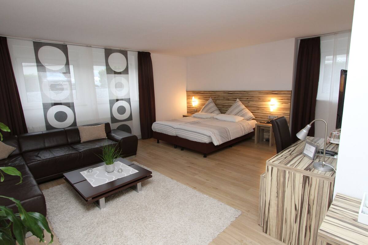 A modern hotel room with a bed, a sofa, a coffee table, and a desk with a lamp.