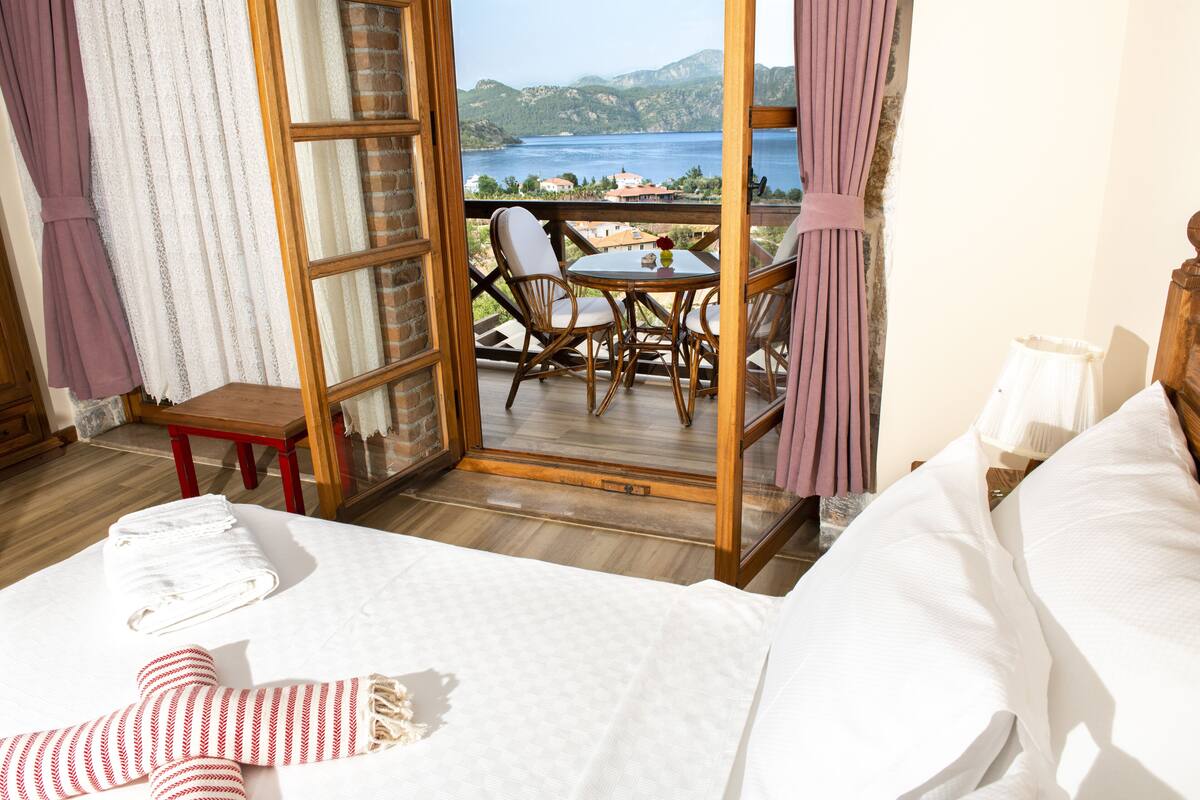 Quarto de hotel com uma cama, uma mesinha com cadeiras e vista para um lago através de uma porta de vidro.
