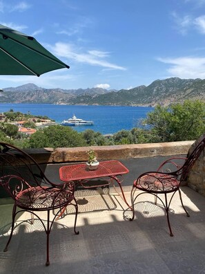 Un patio con dos sillas, una mesa y un paraguas, con vistas a la costa, montañas y un yate.