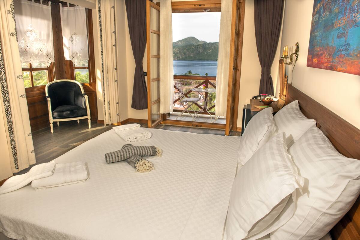 Quarto com cama grande, uma cadeira, vista para um lago e varanda com mobiliário externo.