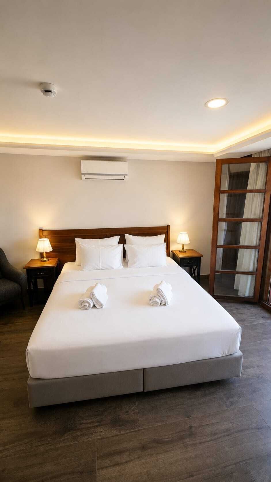 A hotel room with a large bed, two bedside tables with lamps, an air conditioning unit, and a wooden floor.