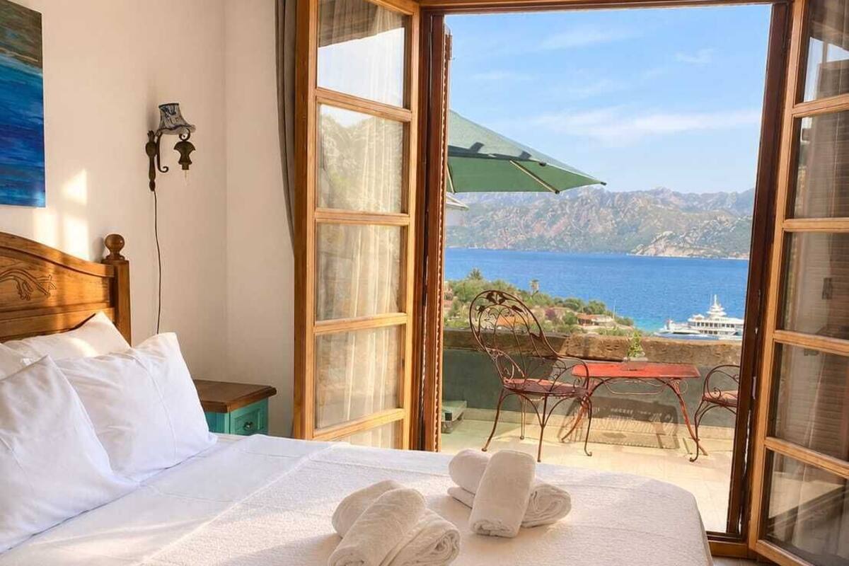 Quarto de hotel com cama, vista para o mar e mesa com cadeiras na área externa.