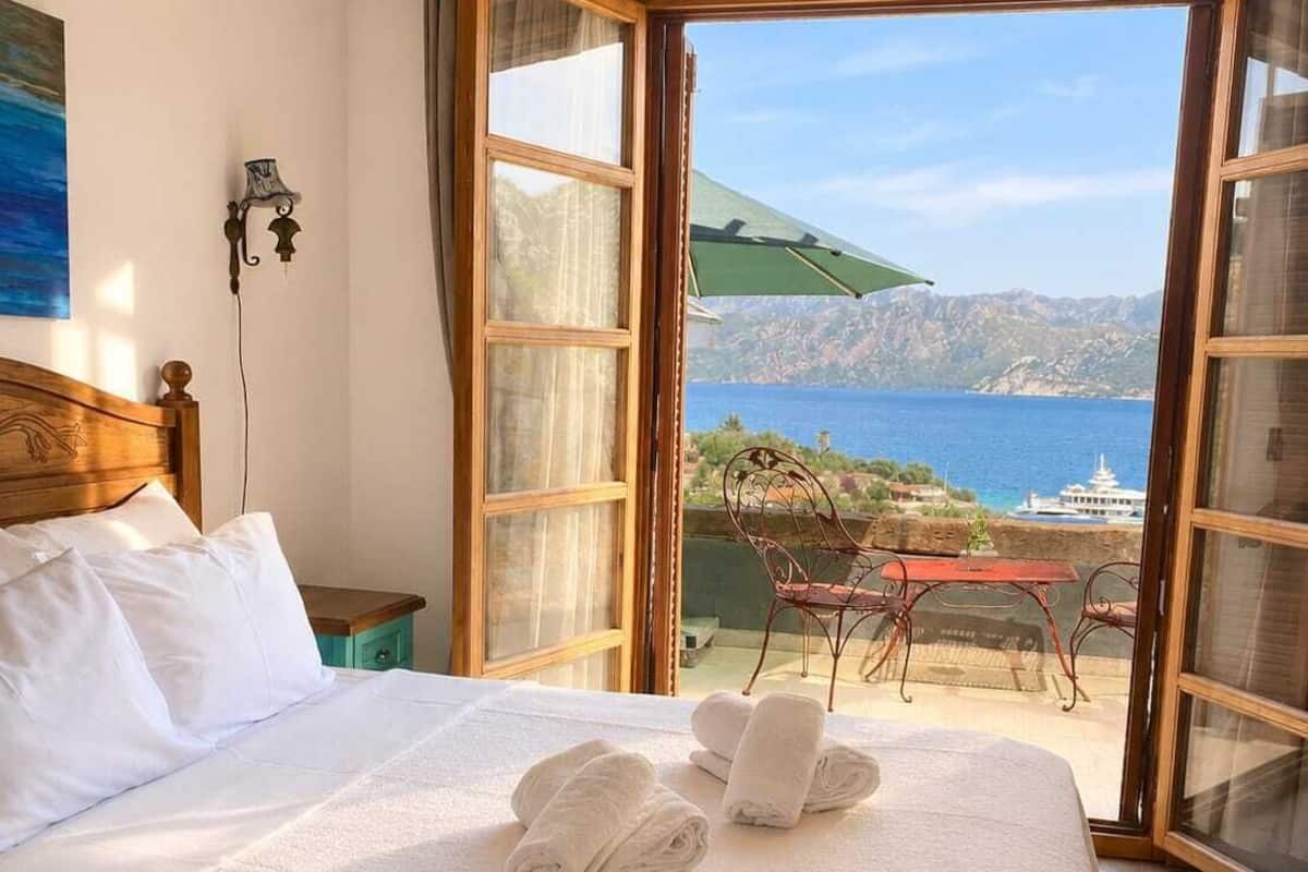 Quarto de hotel com cama, vista para o mar e mesa com cadeiras na área externa.