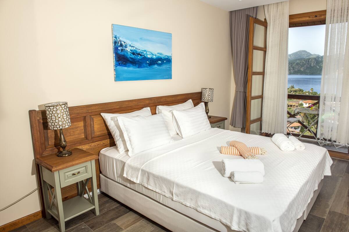 Quarto de hotel com uma cama grande, duas mesas de cabeceira, um quadro na parede e janela com vista para o mar e montanhas.