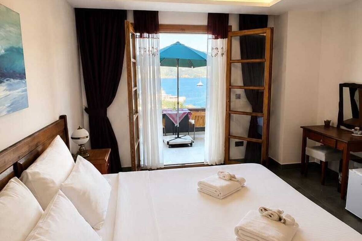 Quarto de hotel com cama, escrivaninha, cadeira, janela com cortinas e vista para o mar.