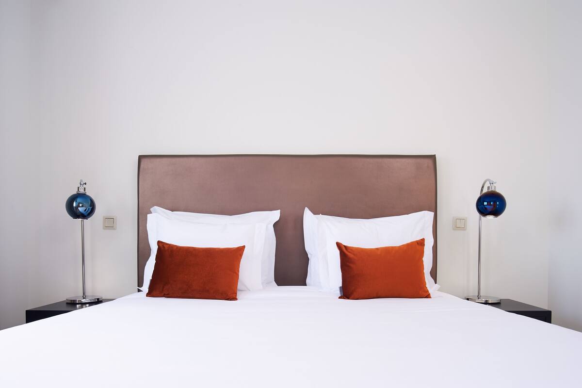 A neatly made bed with white and orange pillows, a brown headboard, and two modern lamps on either side.