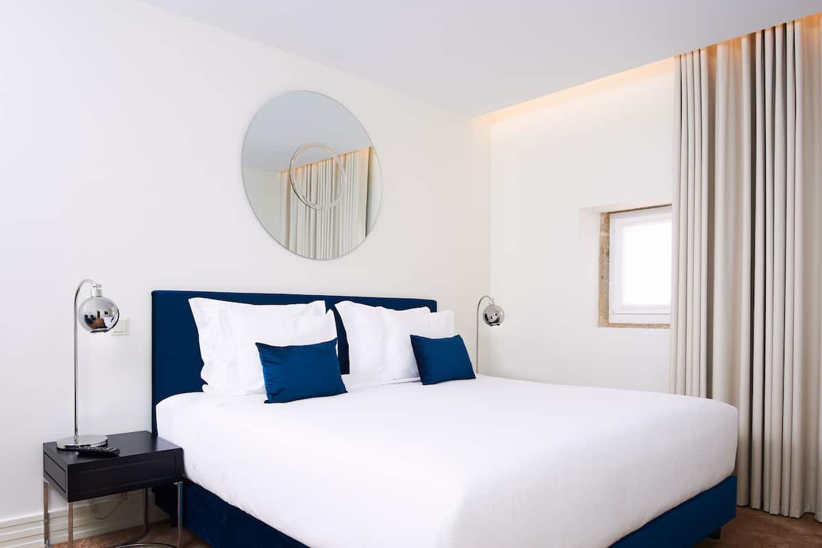 A neatly arranged hotel room with a large bed, bedside lamps, a round mirror, and a window with curtains.