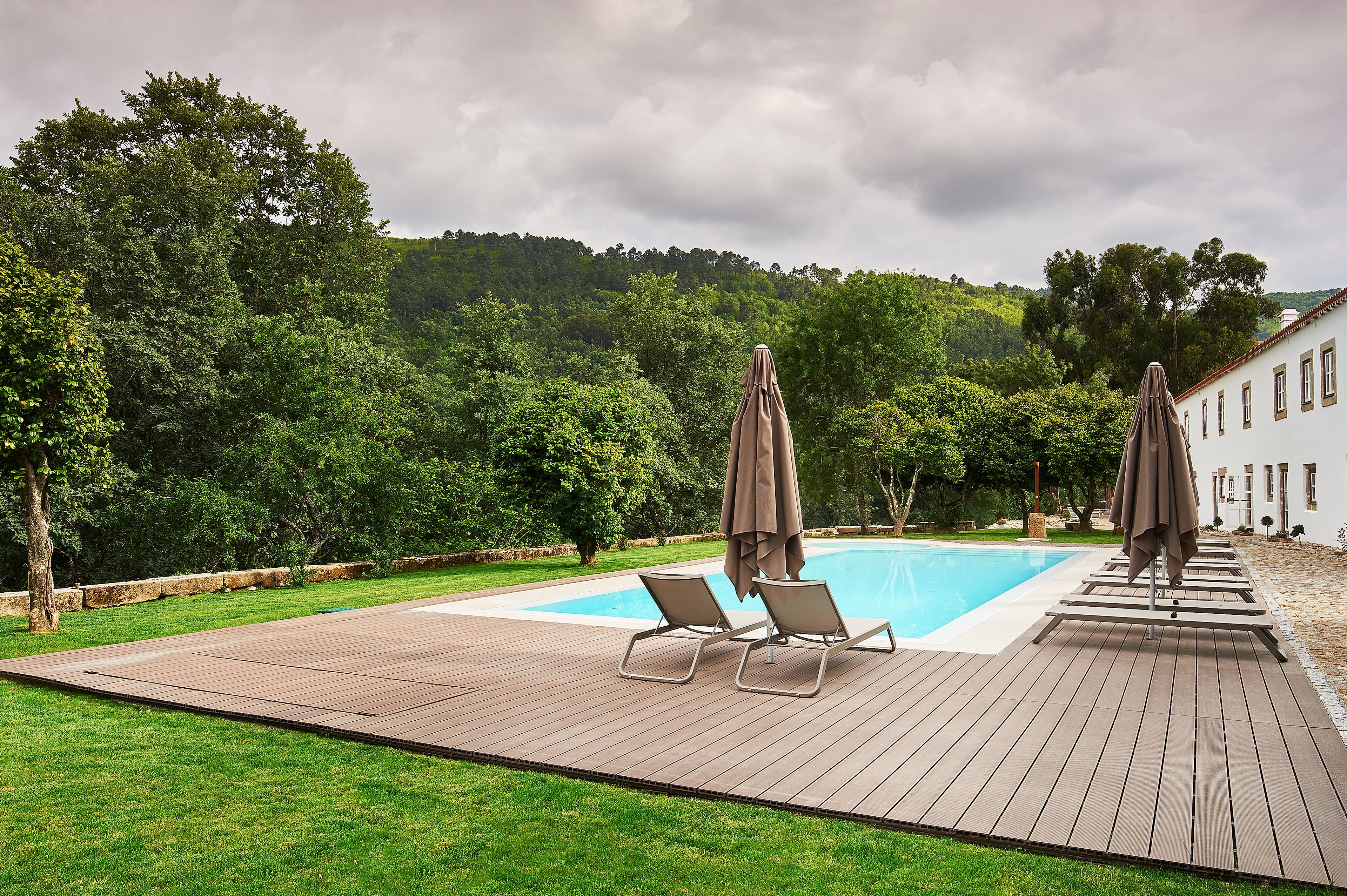 A wooden deck with two lounge chairs and umbrellas, a swimming pool, and a building in the background.