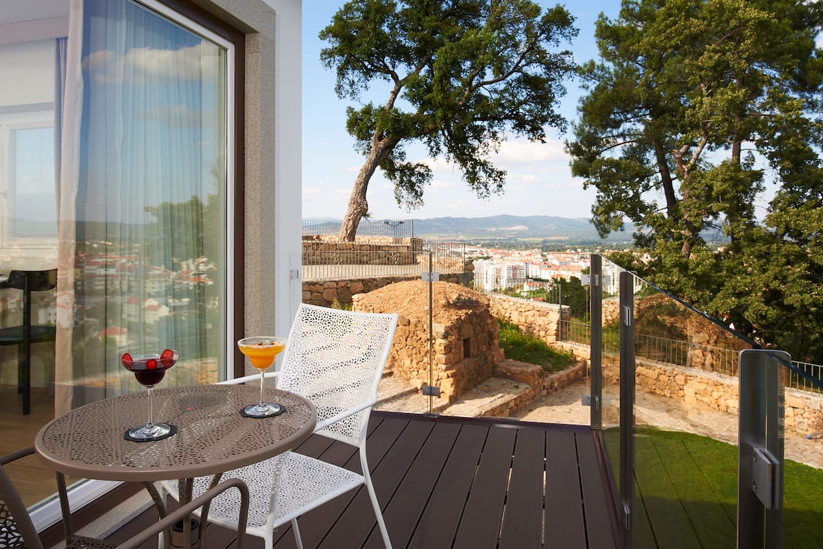 A balcony with a table and chairs, a view of a historic stone wall, and a tree.