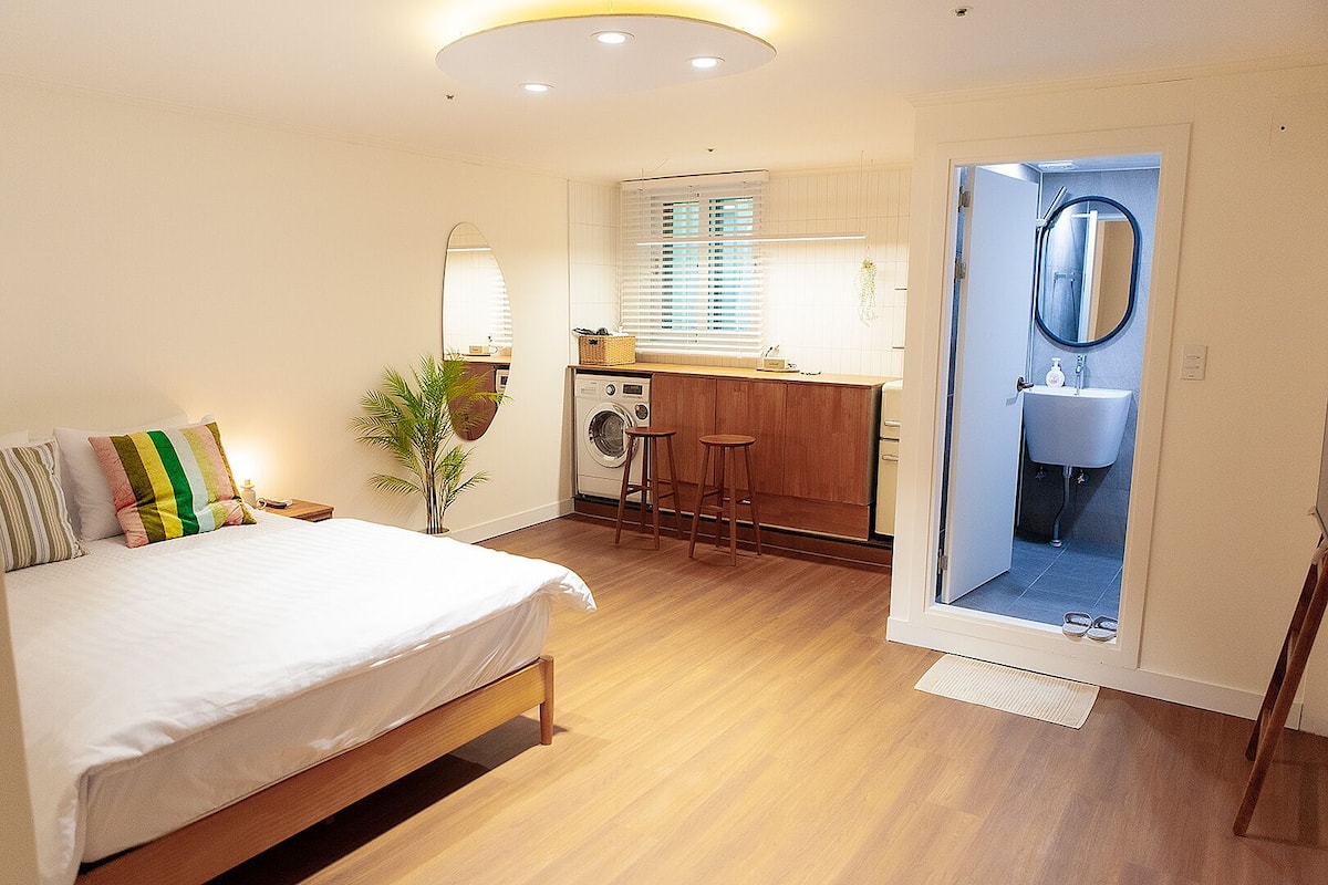 Une chambre d’hôtel compacte comprenant un lit, une petite kitchenette, un lave-linge et une salle de bain avec un miroir et un lavabo.