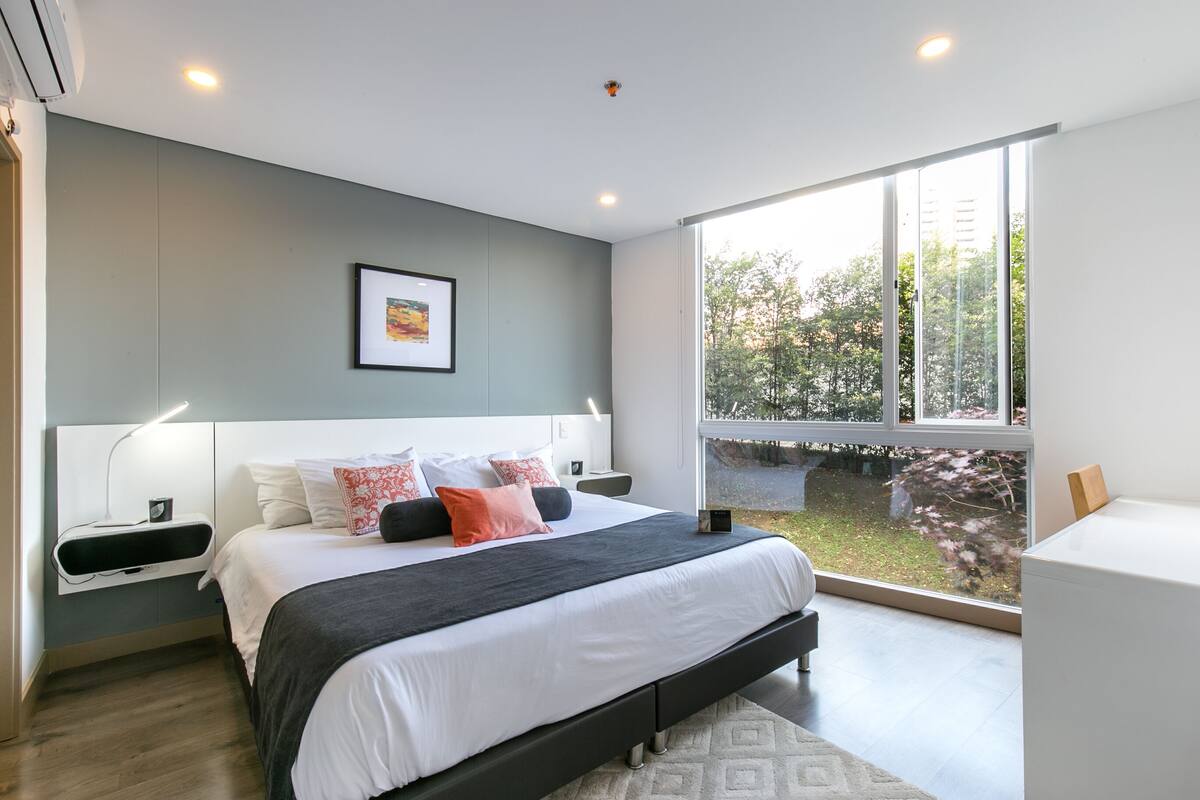 A modern bedroom with a large bed, bedside tables, a nightstand, a window with a view of greenery, and a ceiling light.