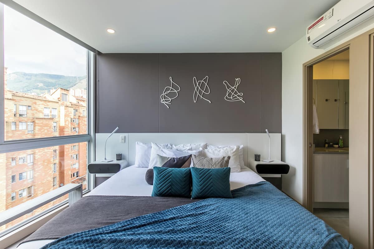 A modern hotel room with a large window, a bed with multiple pillows, a bedside table with a lamp, and a wall-mounted air conditioner.