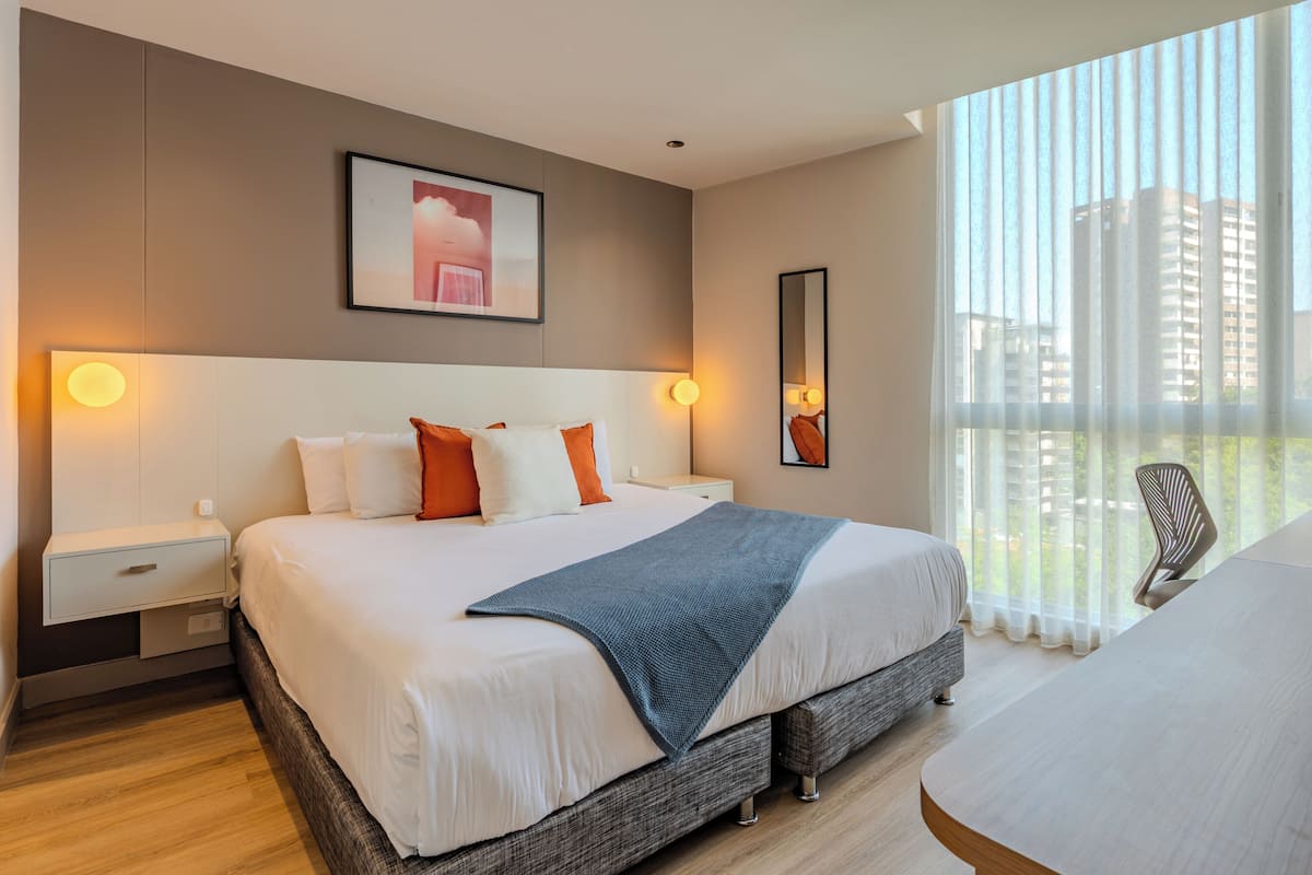 A hotel room with a large bed, bedside tables, a desk, a chair, a window with a view, and a framed picture on the wall.