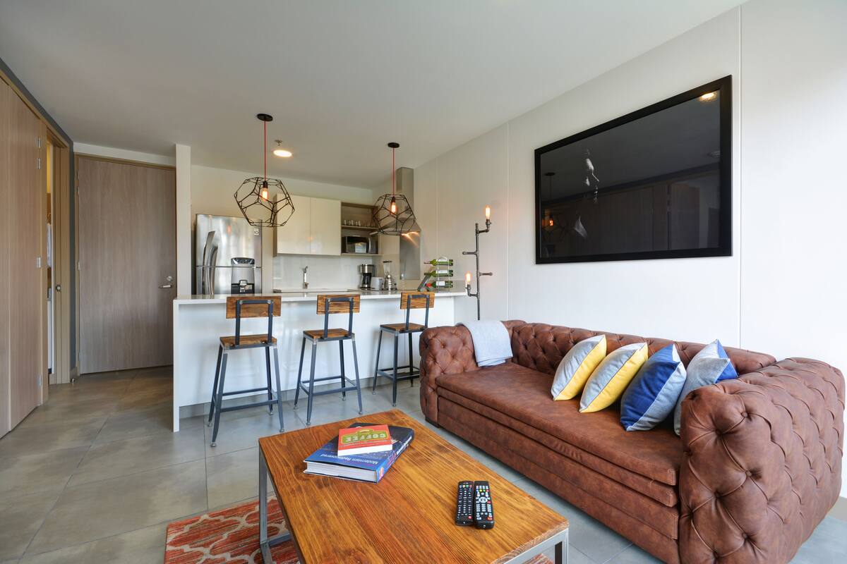 A modern living room with a brown leather sofa, a wooden coffee table with books and remotes, and a kitchen in the background.
