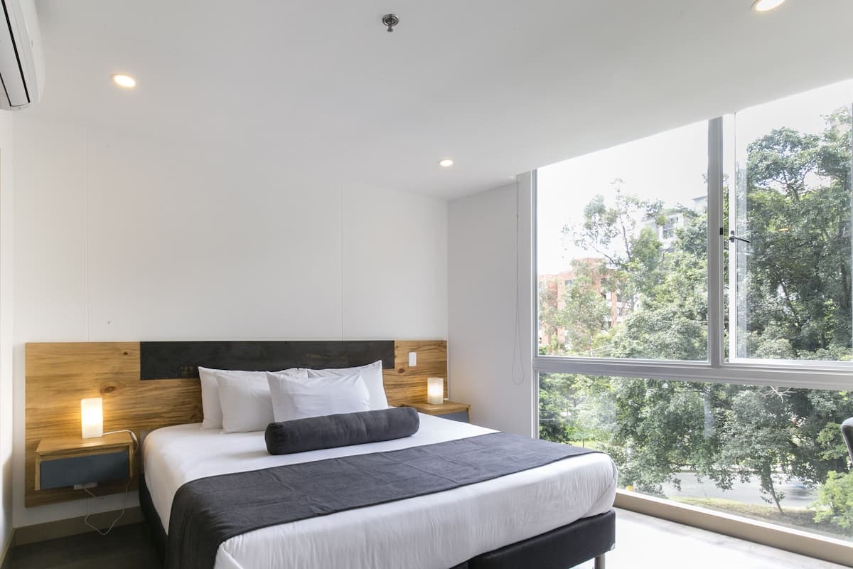 A modern bedroom with a large bed, wooden headboard, bedside tables with lamps, and a large window offering a view of trees and buildings.