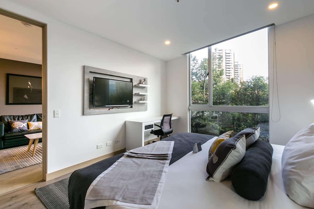 A modern hotel room with a large bed, a desk, a TV mounted on the wall, and a view of the city through the window.