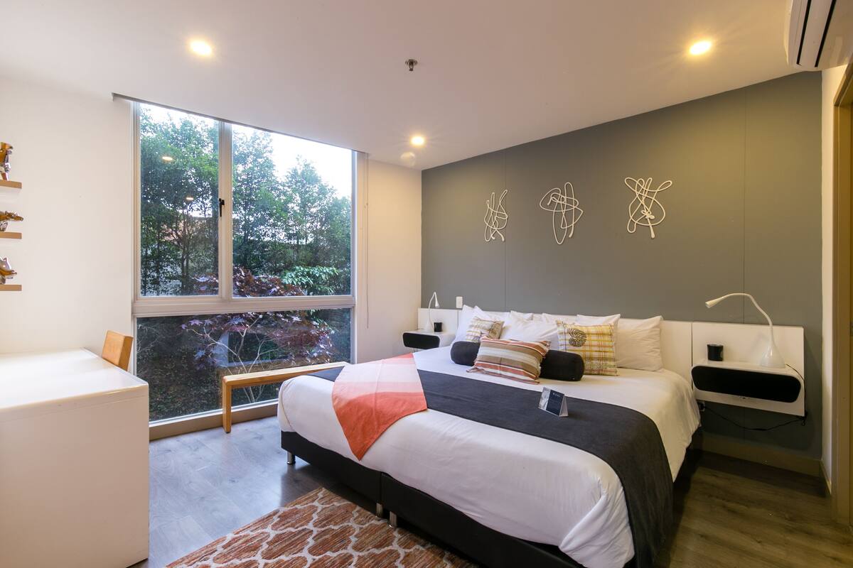 A modern bedroom with a large window, a bed with multiple pillows, bedside tables, and abstract wall art.