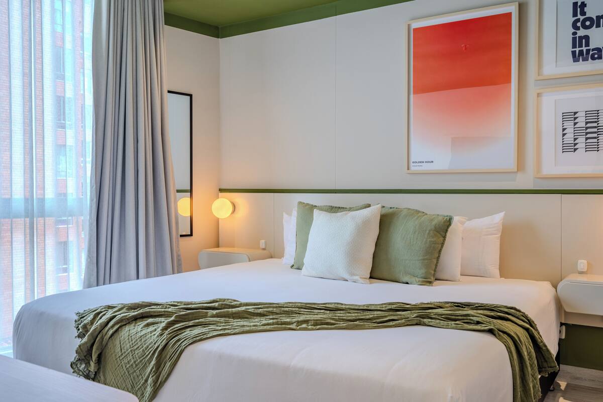 A hotel room with a bed, two bedside tables, a wall-mounted light, a window with curtains, and a framed artwork above the bed.