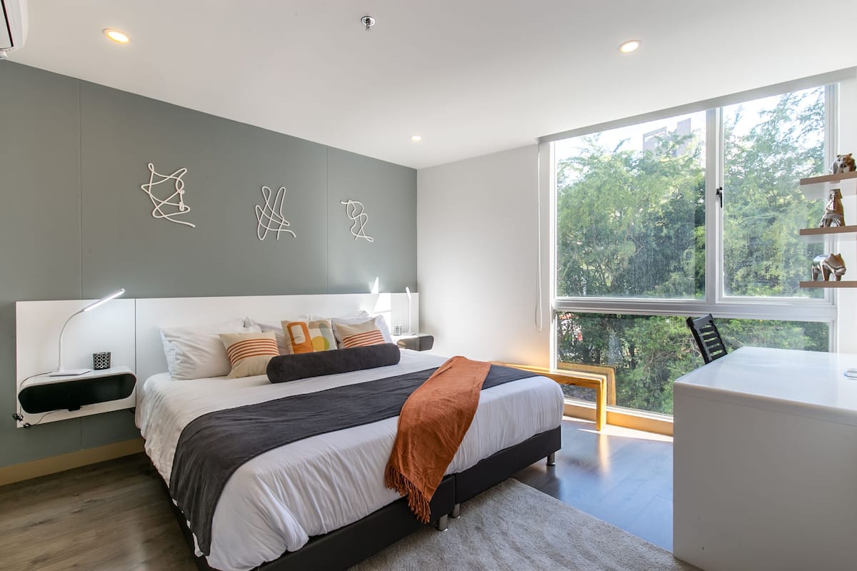 A modern bedroom with a large bed, bedside tables, a desk, and a window with a view of trees.