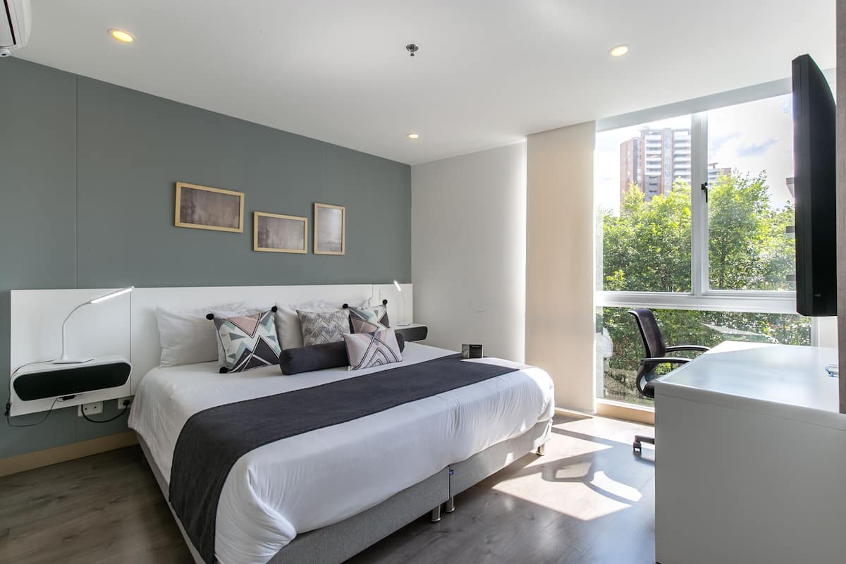 A modern bedroom with a large bed, bedside tables, a desk, and a window with a view of buildings and trees.