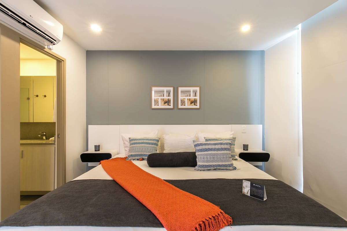 A modern hotel room with a large bed, a bedside table, a nightstand, and a wall-mounted air conditioner.