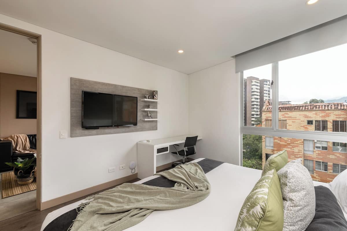 A modern hotel room with a large bed, a desk, a TV mounted on the wall, and a view of the city through the window.