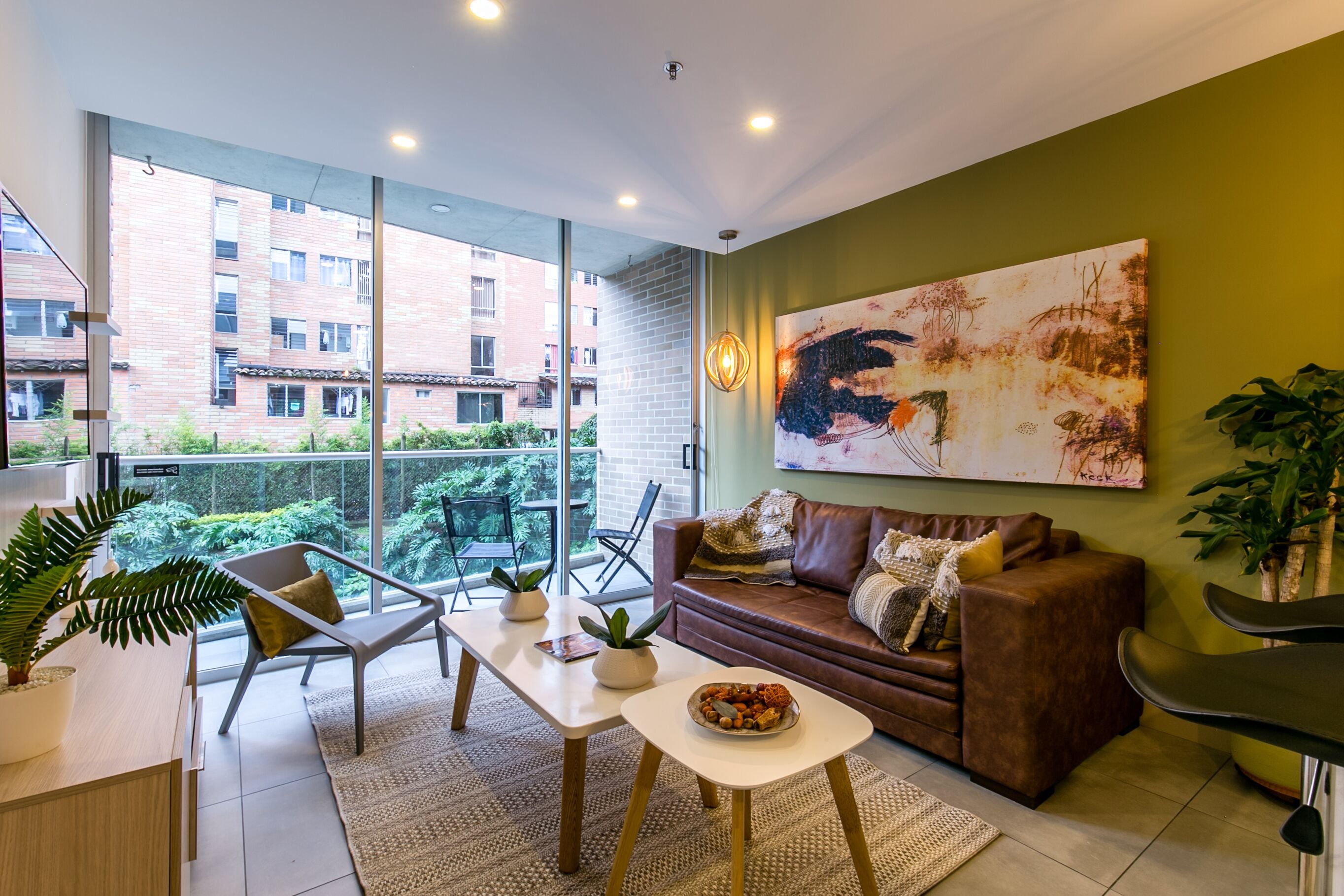 A modern living room with a brown leather sofa, a white coffee table, and a large abstract painting on the wall. There are two potted plants, a glass table, and a view of a brick building through large windows.