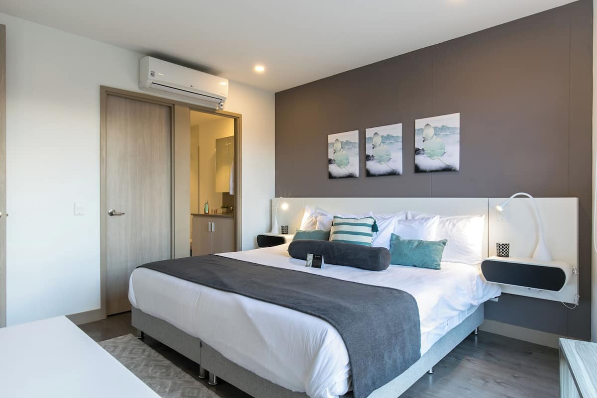A modern bedroom with a large bed, bedside tables, a wall-mounted air conditioner, and artwork.