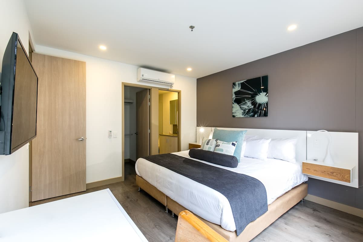 A modern bedroom with a large bed, a bedside table, an air conditioning unit, and a door leading to another room.