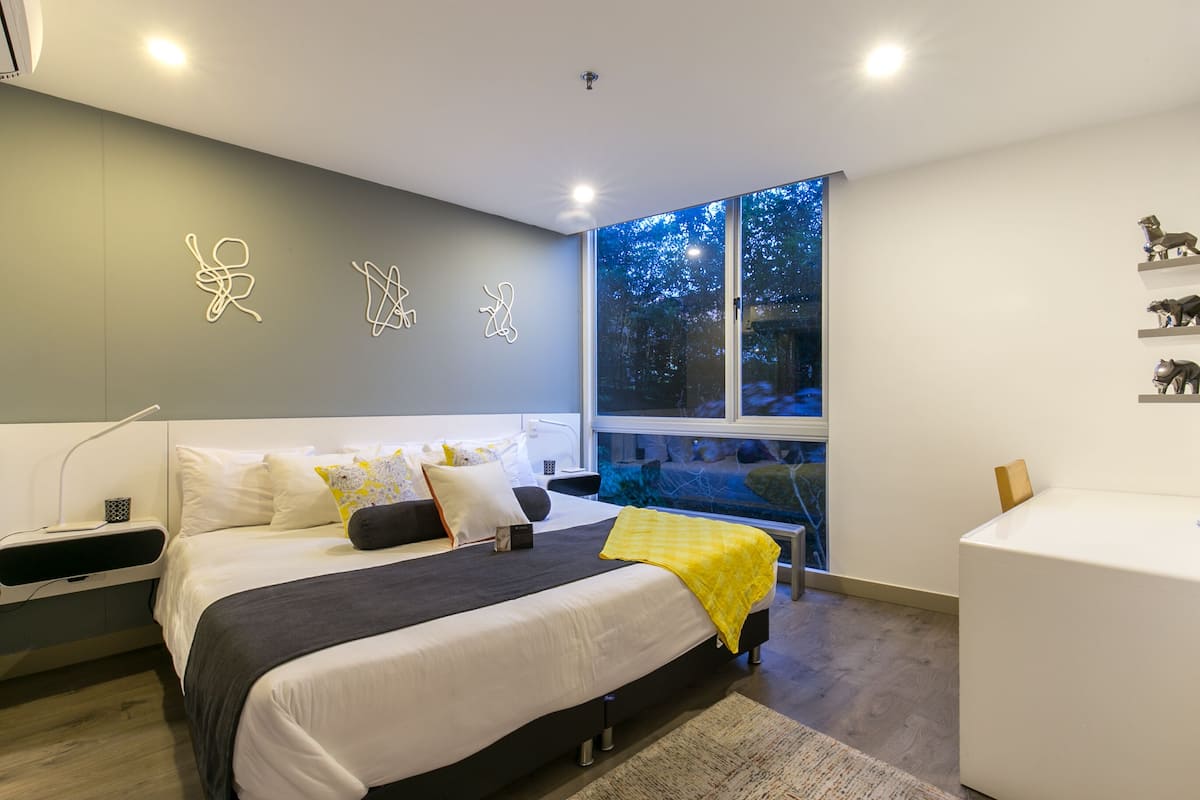 A modern bedroom with a large bed, bedside tables, a desk, and a window with a view of trees.