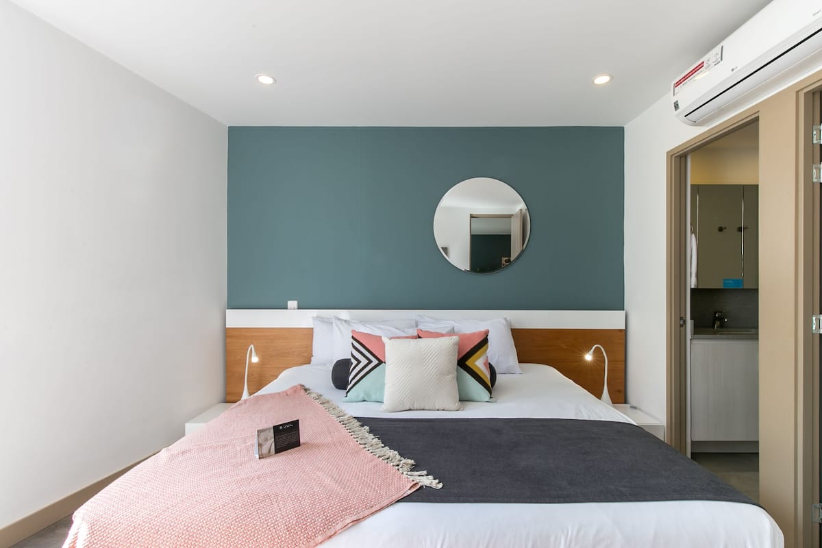 A modern hotel room with a neatly made bed, a round mirror on the wall, and a view into the adjacent kitchen area.