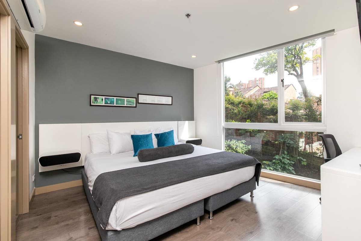 A modern bedroom with a large bed, a desk, and a window with a view of greenery and buildings.