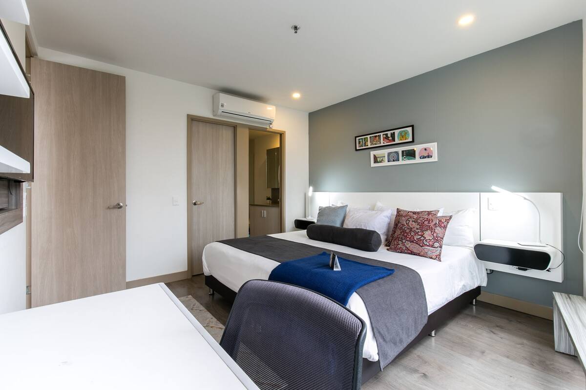 A modern bedroom with a bed, bedside tables, a desk, and an air conditioning unit.