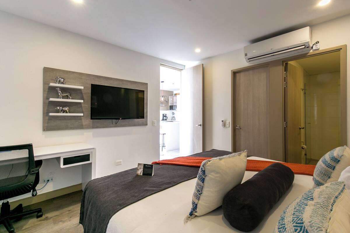 A modern bedroom with a bed, a desk, a TV mounted on the wall, and an air conditioning unit.