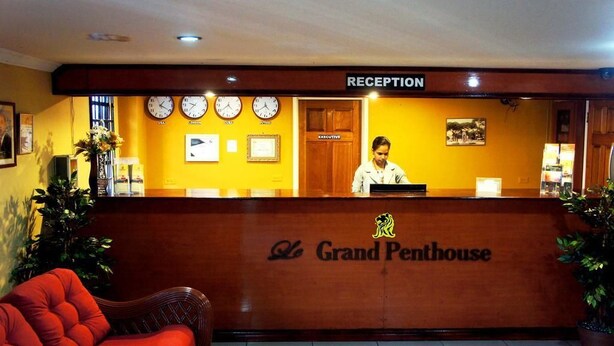 Mostrador de recepción en el hotel Grand Penthouse, con relojes, una recepcionista y una silla roja.