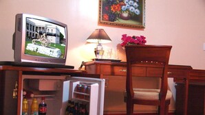 Una habitación con una televisión sobre un mueble de madera, un refrigerador pequeño con bebidas, una silla de madera y un cuadro de flores en la pared.