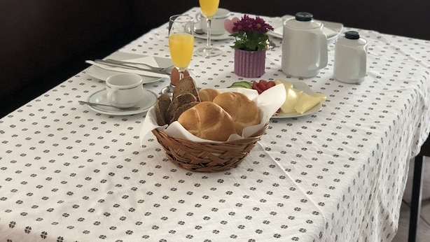 Une table à manger recouverte d’une nappe blanche, un panier de pain, une carafe, une petite plante en pot et deux verres de jus d’orange.