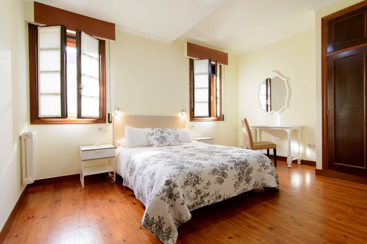 A bedroom with a wooden floor, a bed with a floral bedspread, a nightstand, a chair, a small table, a mirror, and a door.