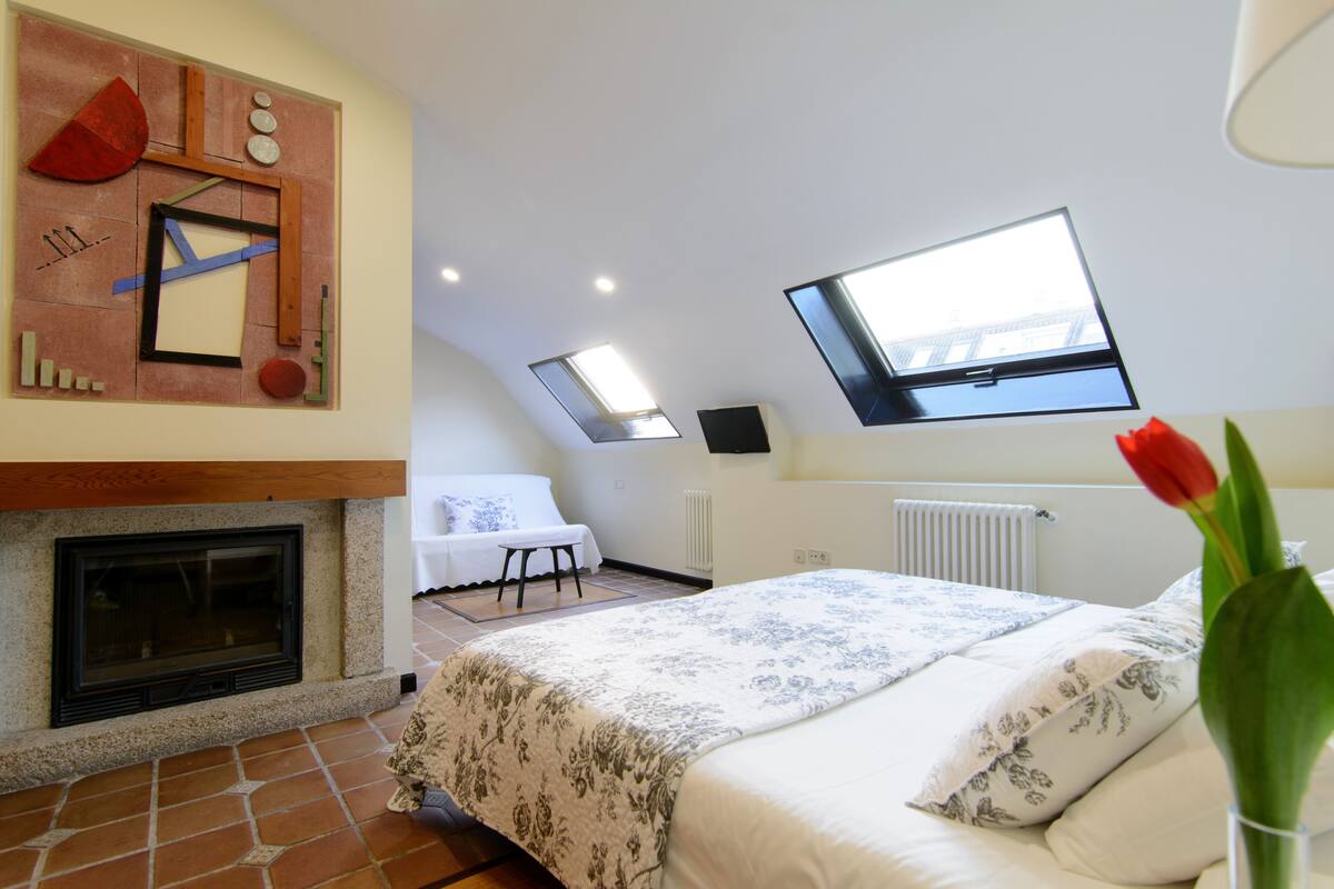 A bedroom with a fireplace, a bed with floral bedding, a TV mounted on the wall, and a skylight.
