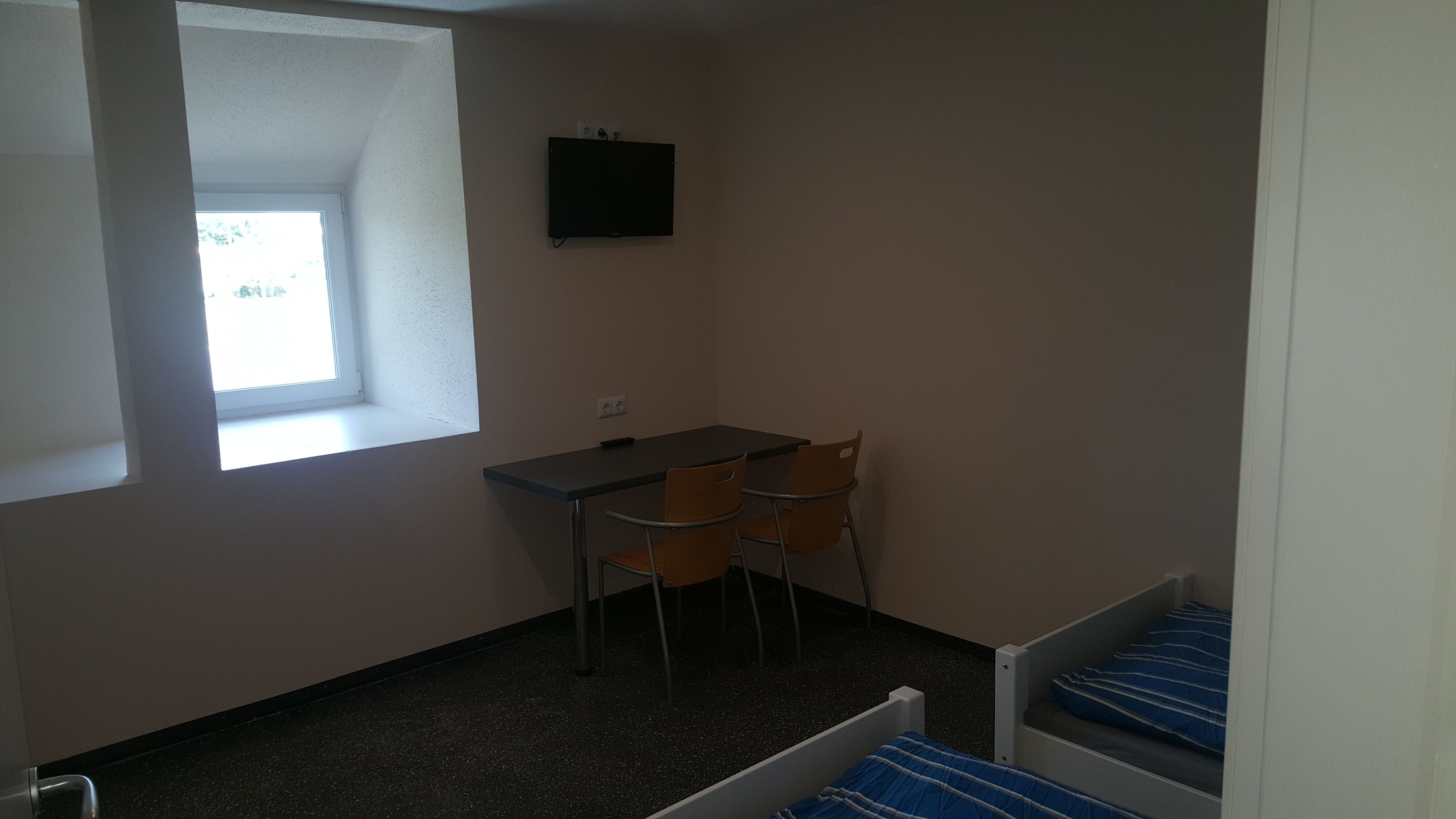 Ein kleines Zimmer mit Schreibtisch, zwei Stühlen, einem Fenster, einem Bett und einem am Wand befestigten Fernseher.