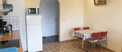 Eine kleine Küche mit Kühlschrank, Mikrowelle und Kaffeemaschine. Ein Esstisch mit Stühlen und ein Fliesenboden sind vorhanden.