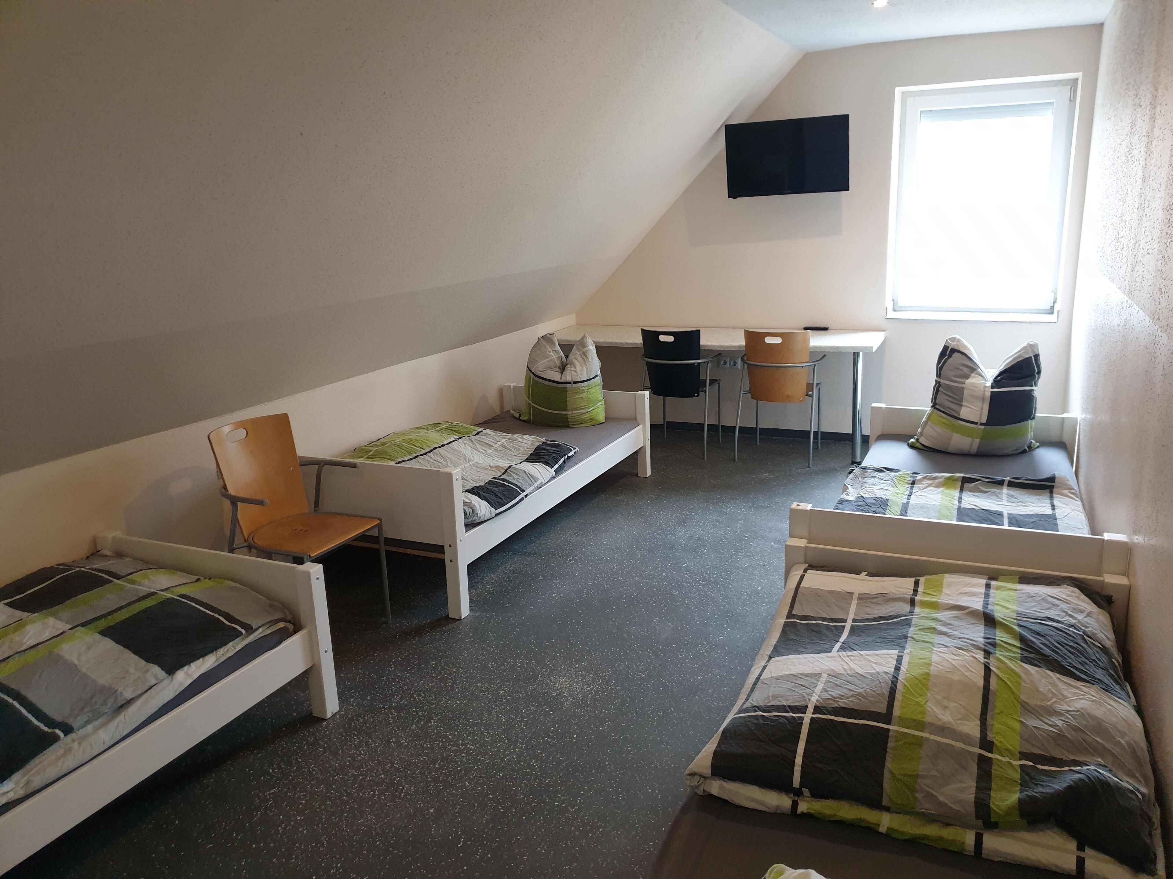Ein Schlafsaalzimmer mit zwei Einzelbetten, einem Schreibtisch, zwei Stühlen und einem an der Wand befestigten Fernseher.
