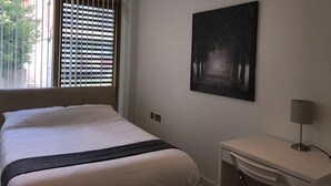 A hotel room with a bed, a desk, a lamp, and a painting of a forest path.