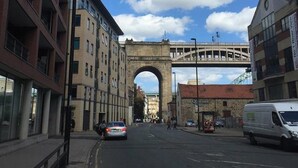 A street view with a large stone arch bridge, buildings on either side, and a white van parked on the right.
