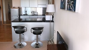 A modern kitchen with white cabinets, a black countertop, and two bar stools. There is a microwave, a kettle, and a coffee maker on the counter.